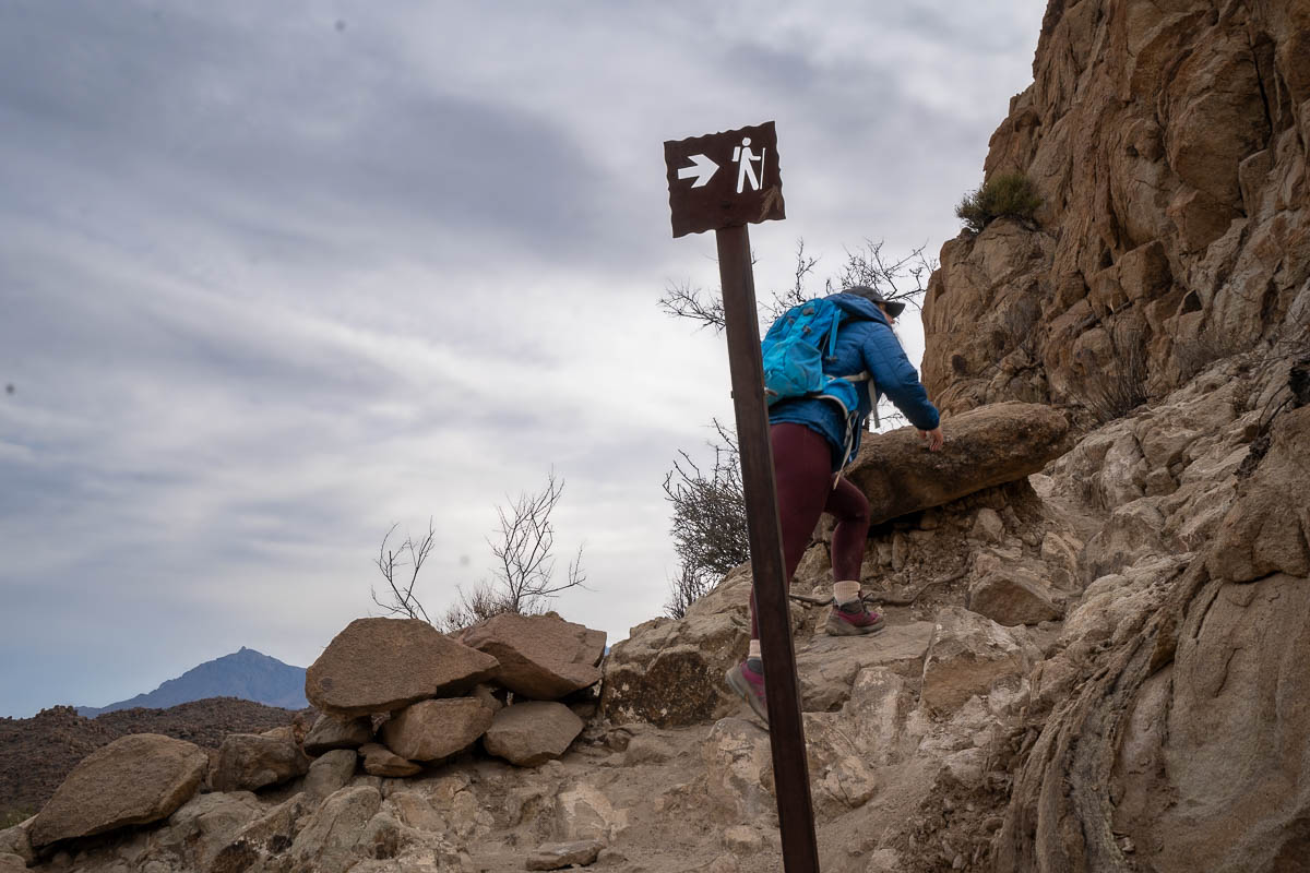 Woman hiking past a metal sign stuck into some boulders along the Balanced Rock Trail in Big Bend National Park in Texas