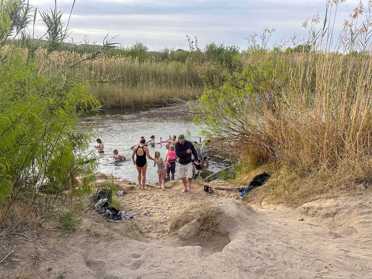 Crowd of people sitting in and walking to Big Bend Hot Springs with reeds around it in Big Bend National Park in Texas