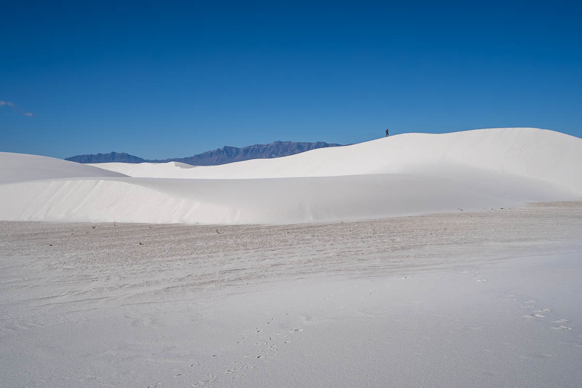 Woman walking across a white sand dune with mountains in the background along the Alkali Flat Trail in White Sands National Park in New Mexico