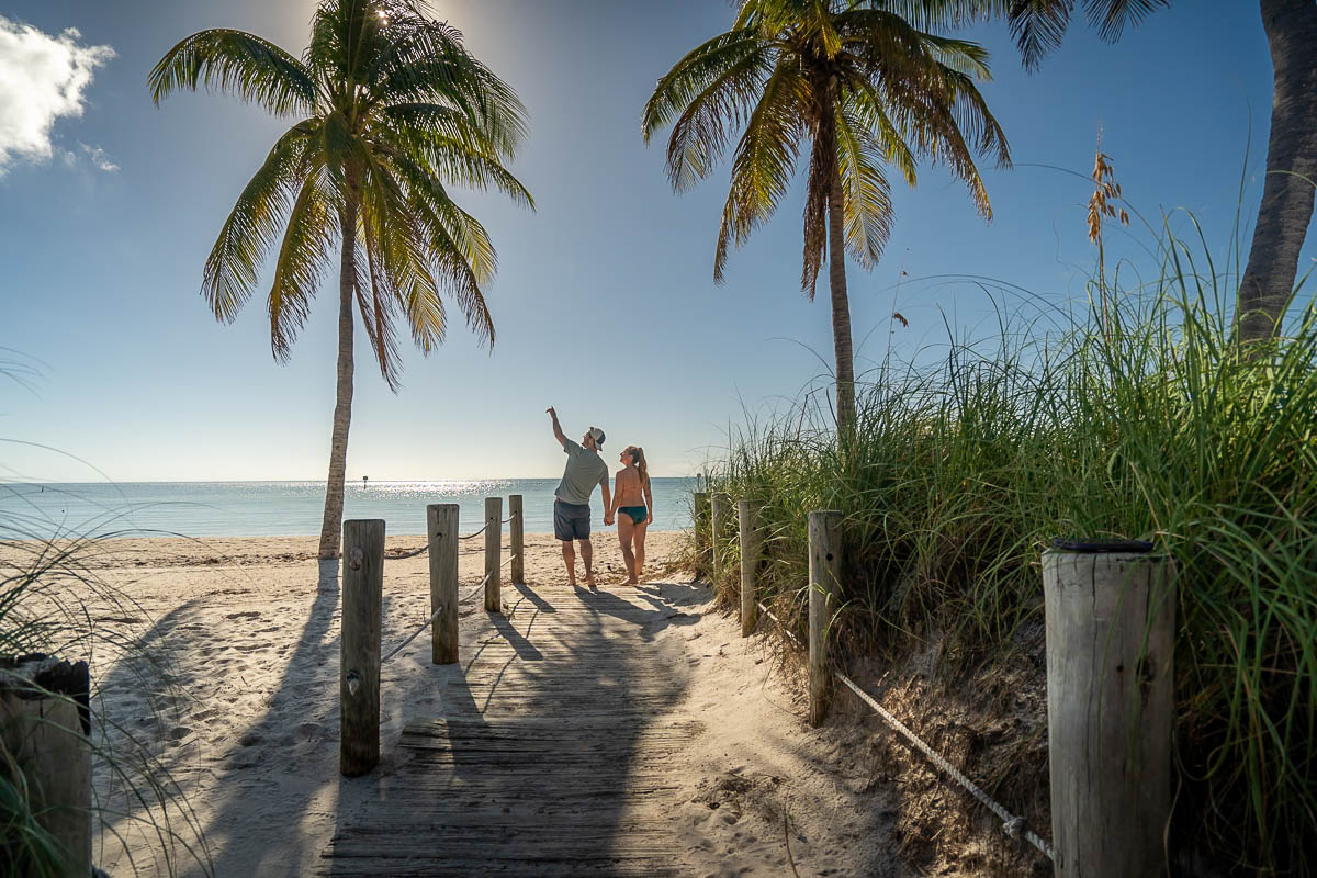 Couple holding hands and walking along a wooden boardwalk between two palm trees at Smathers Beach in Key West, Florida