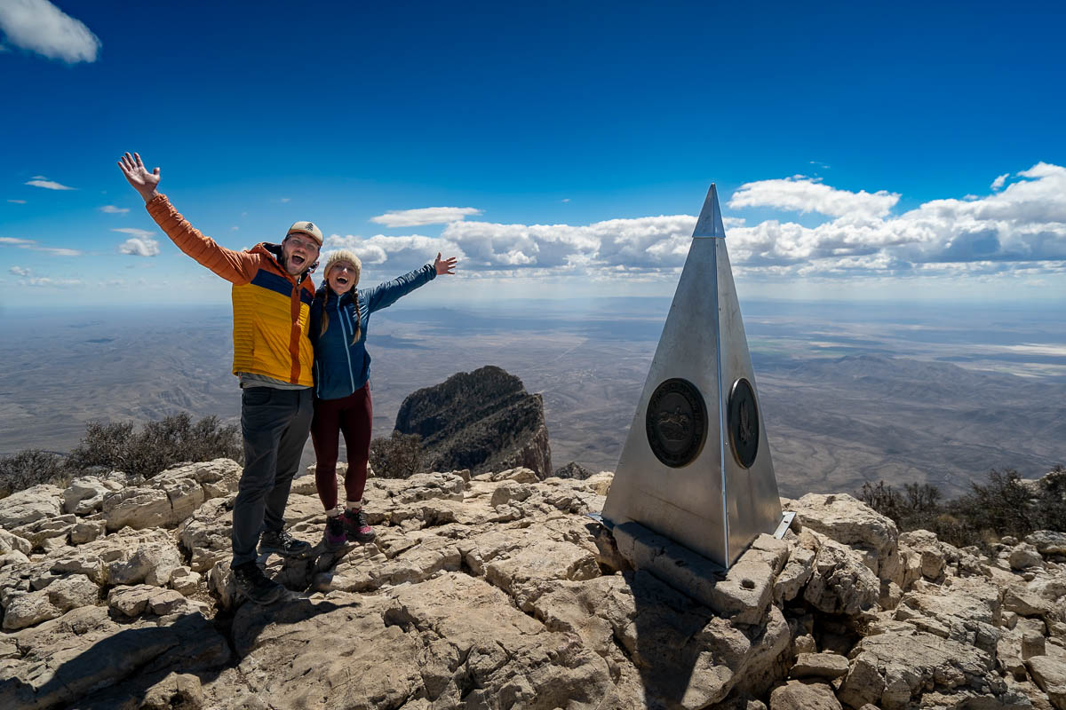 Couple posing next to a silver obelisk at the top of Guadalupe Peak with El Capitan in the background in Guadalupe Mountains National Park in Texas