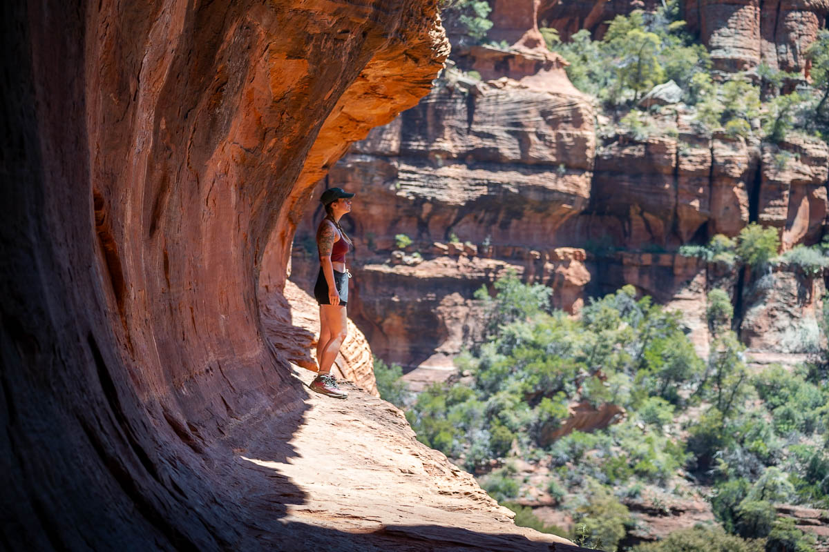 Woman standing on a ledge of the Subway Cave with rock formations and greenery in the background in Sedona, Arizona