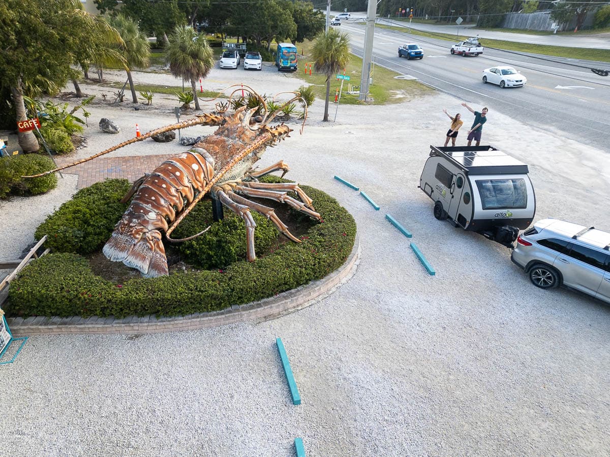 Couple standing on the roof of the Safari Condo Alto F1743 Expedition trailer next to Betsy the Lobster in Rain Barrel Village in Islamorada in the Florida Keys