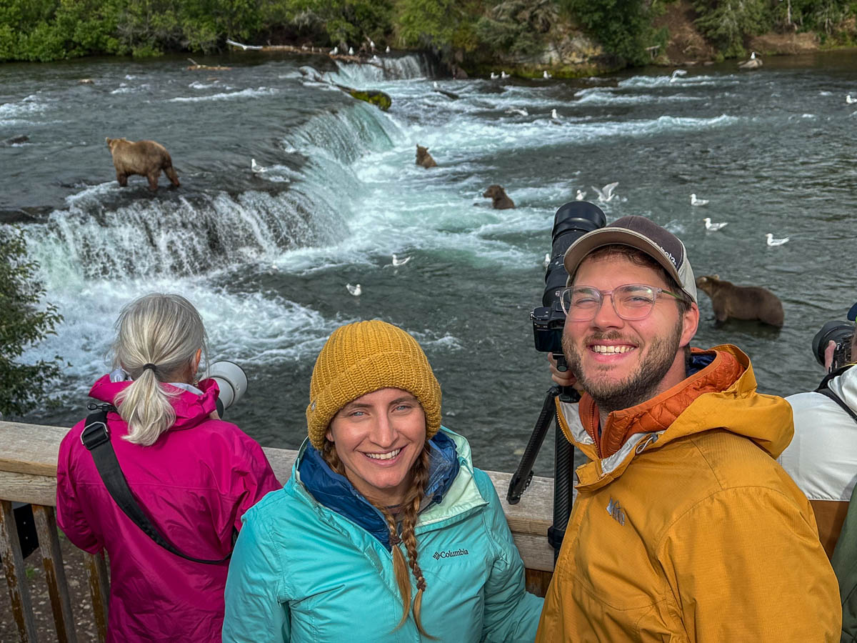 Couple smiling on the Brooks Falls Platform with brown bears in the background of Brooks Falls in Katmai National Park