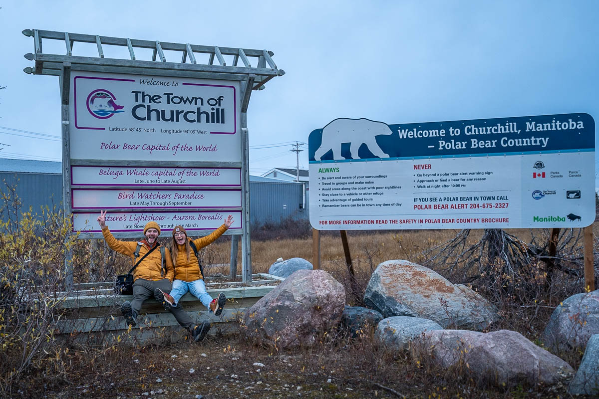 Couple sitting on a sign for Churchill, Manitoba, the polar bear capital of the world