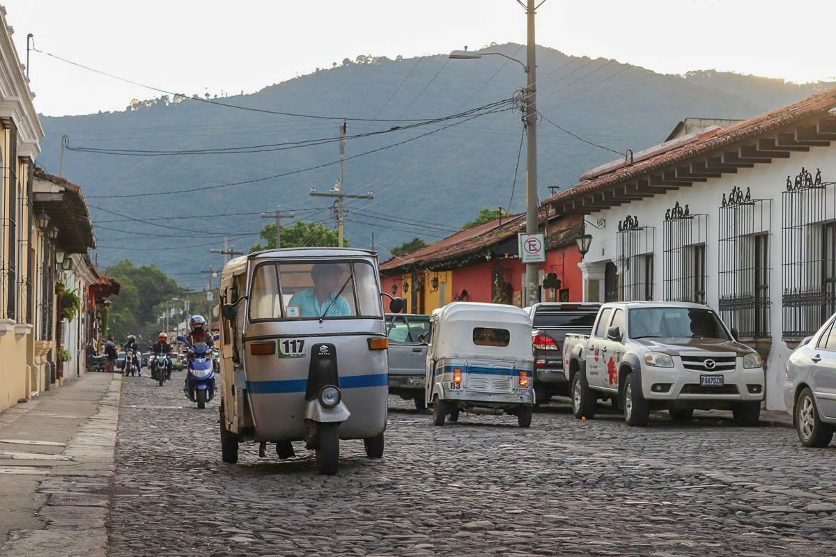 Tuk-tuks driving down a cobblestone street with cars parked in the background in Antigua, Guatemala