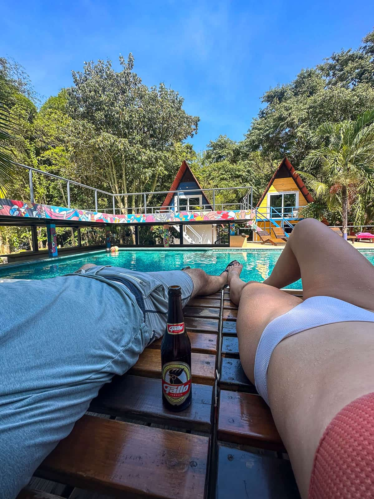 Legs of a couple laying by the pool with A-frame cabins in the background of Greengos Hotel near Semuc Champey, Guatemala