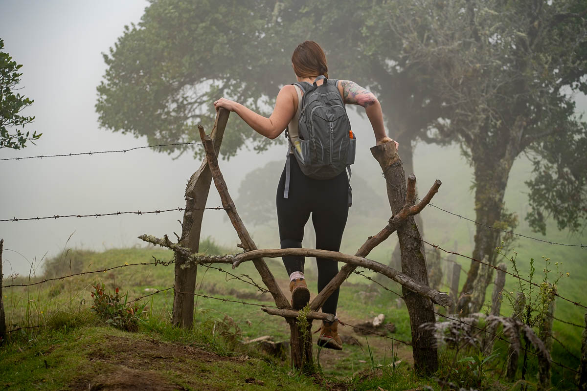 Woman climbing over an opening in a barbed wire fence with clouds in the background along the Cerro Morrogacho trail in the Cocora Valley near Salento, Colombia