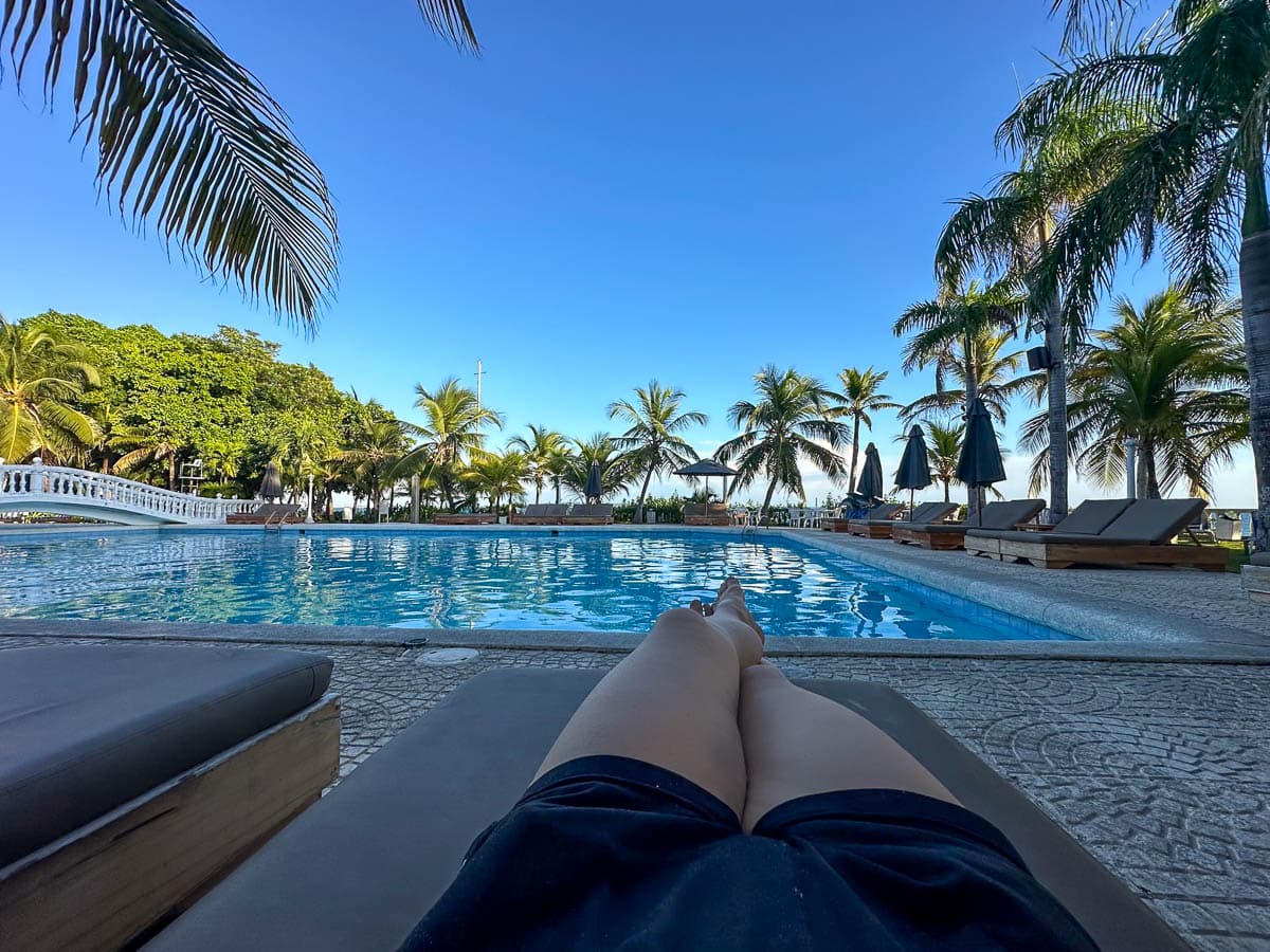 Woman's legs outstretched on a lounge chair in front of a pool and palm trees at a resort on Isla Grande in Colombia