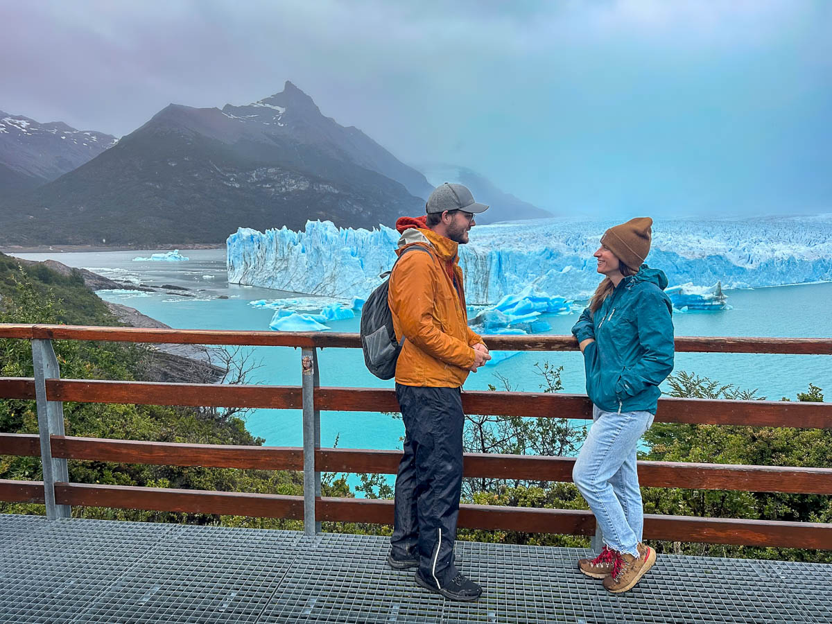 Couple standing by a railing with a Perito Moreno glacier and mountains in the background along the Sendero de la Costa trail in Los Glaciares National Park near El Calafate, Argentina