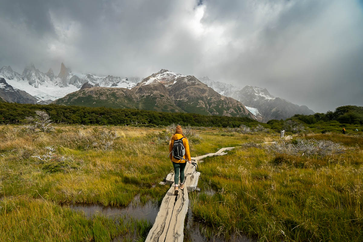 Woman hiking on a rustic wooden bridge through a bog with snowy mountains in the background along the Laguna de los Tres trail in El Chaltén, Argentina