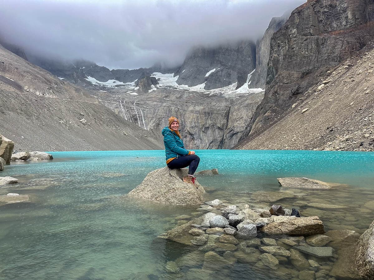Woman sitting on a rock in Lago Torres with mountains covered by clouds in the background along the Mirador Base las Torres Trail in Torres del Paine National Park in Chile
