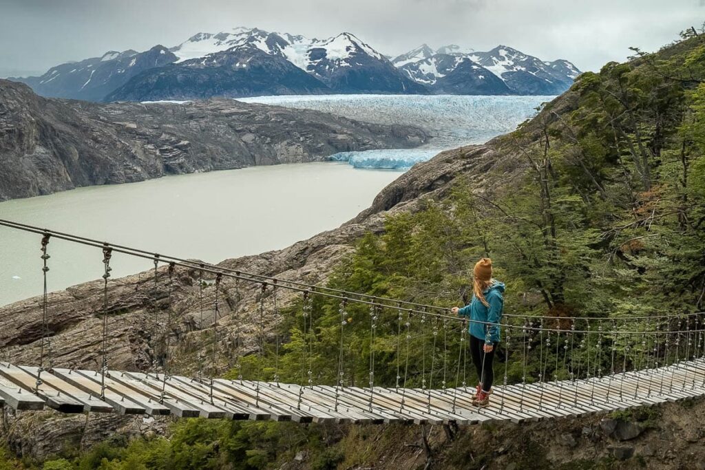 Woman walking across a suspension bridge while looking at Glacier Grey in Torres del Paine National Park in Chilean Patagonia