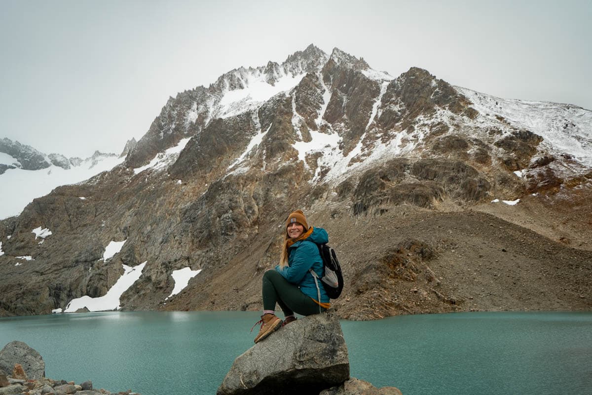 Woman sitting on a rock in front of an alpine lake and snowy mountain along the Laguna de los Tres Trail in Argentinian Patagonia