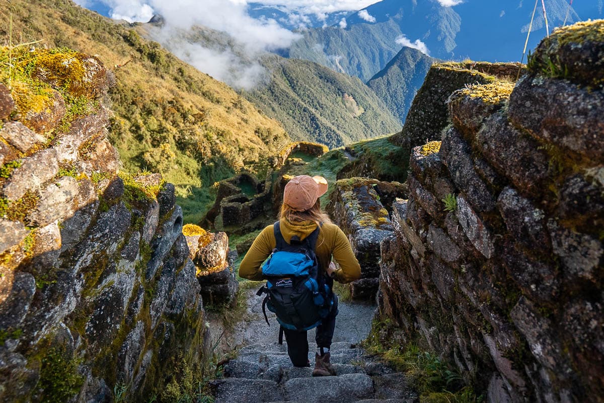 Woman hiking down stairs at Phuyupatamarca ruins with Andes Mountains in the background along the Inca Trail in Peru