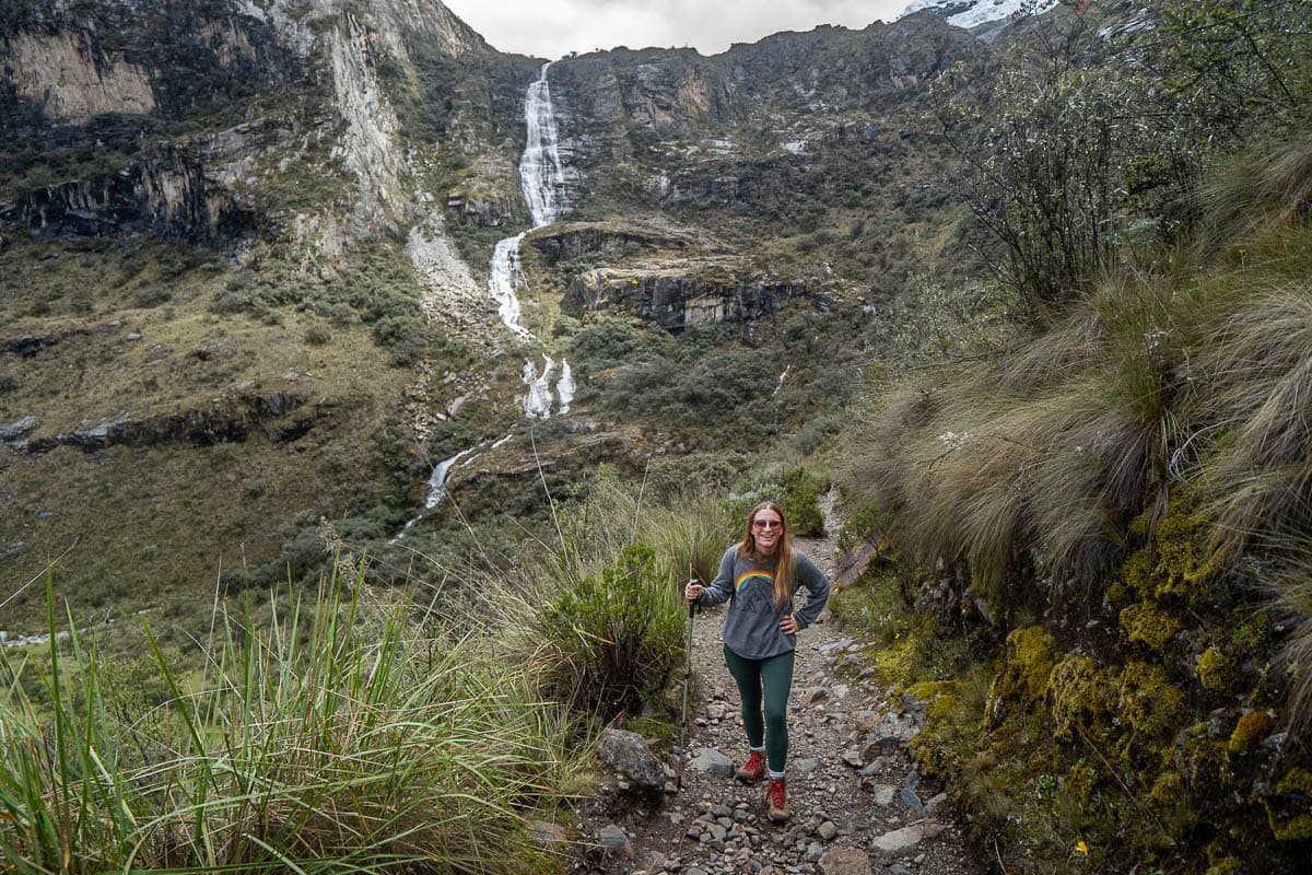 Smiling woman holding a trekking pole with a rugged mountain and waterfall in the background along the Laguna 69 hike in Huascarán National Park in northern Peru