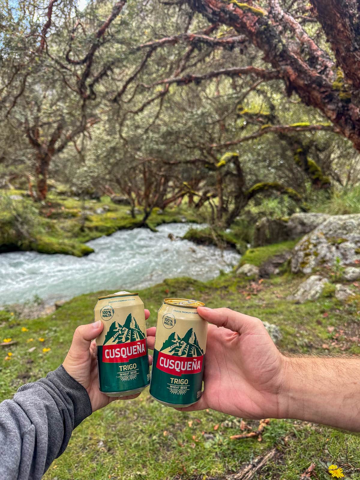 Couple holding beers in front of Rio Ranrahirca and trees in the background along the Laguna 69 hike in Huascarán National Park in northern Peru
