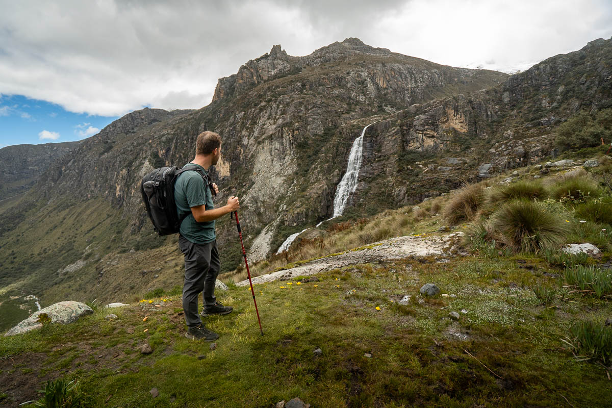 Man standing along the Laguna 69 hike with a rugged cliffside and waterfall in the background in northern Peru