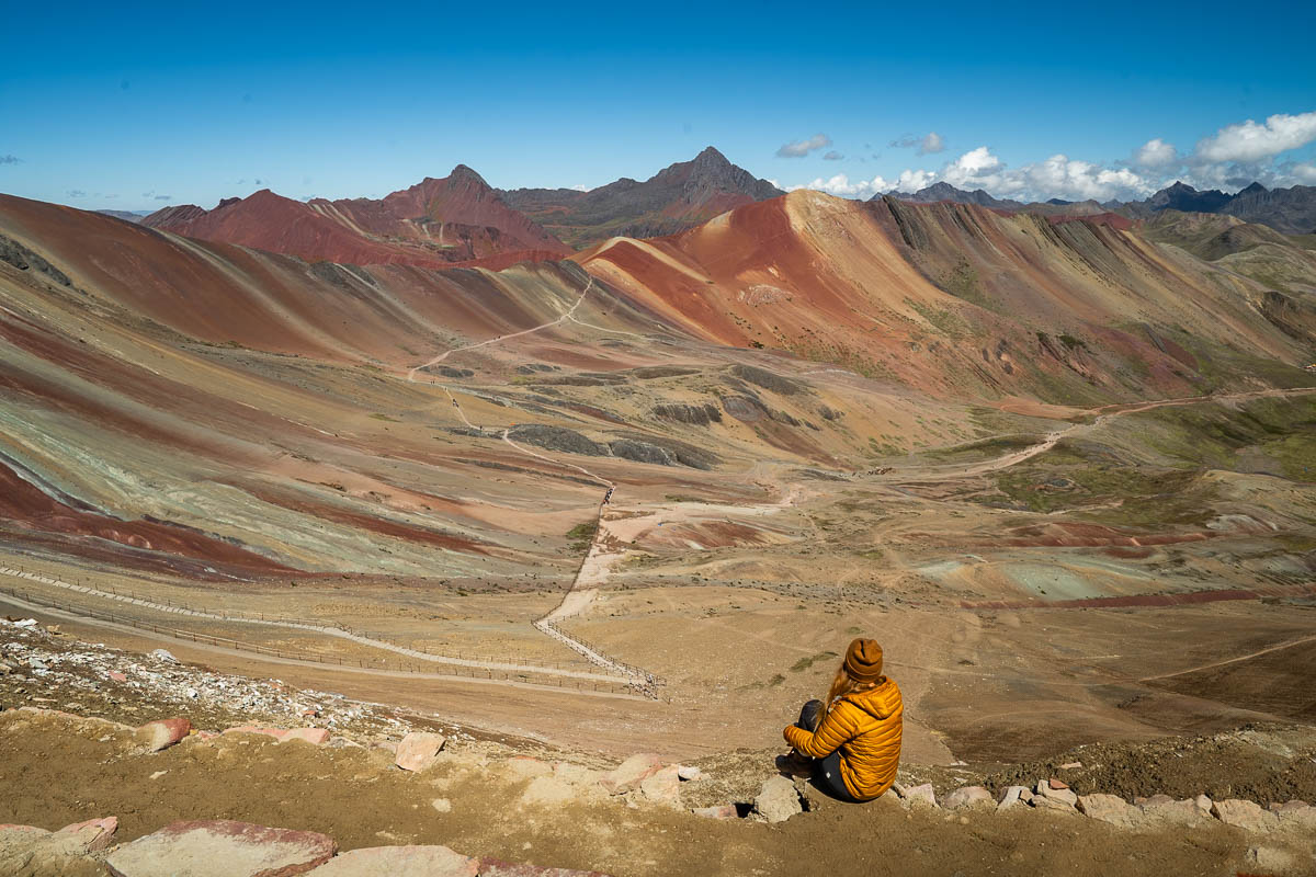 Woman sitting on a cliff looking at the colorful striations of the Red Valley along the Rainbow Mountain hike near Cusco, Peru