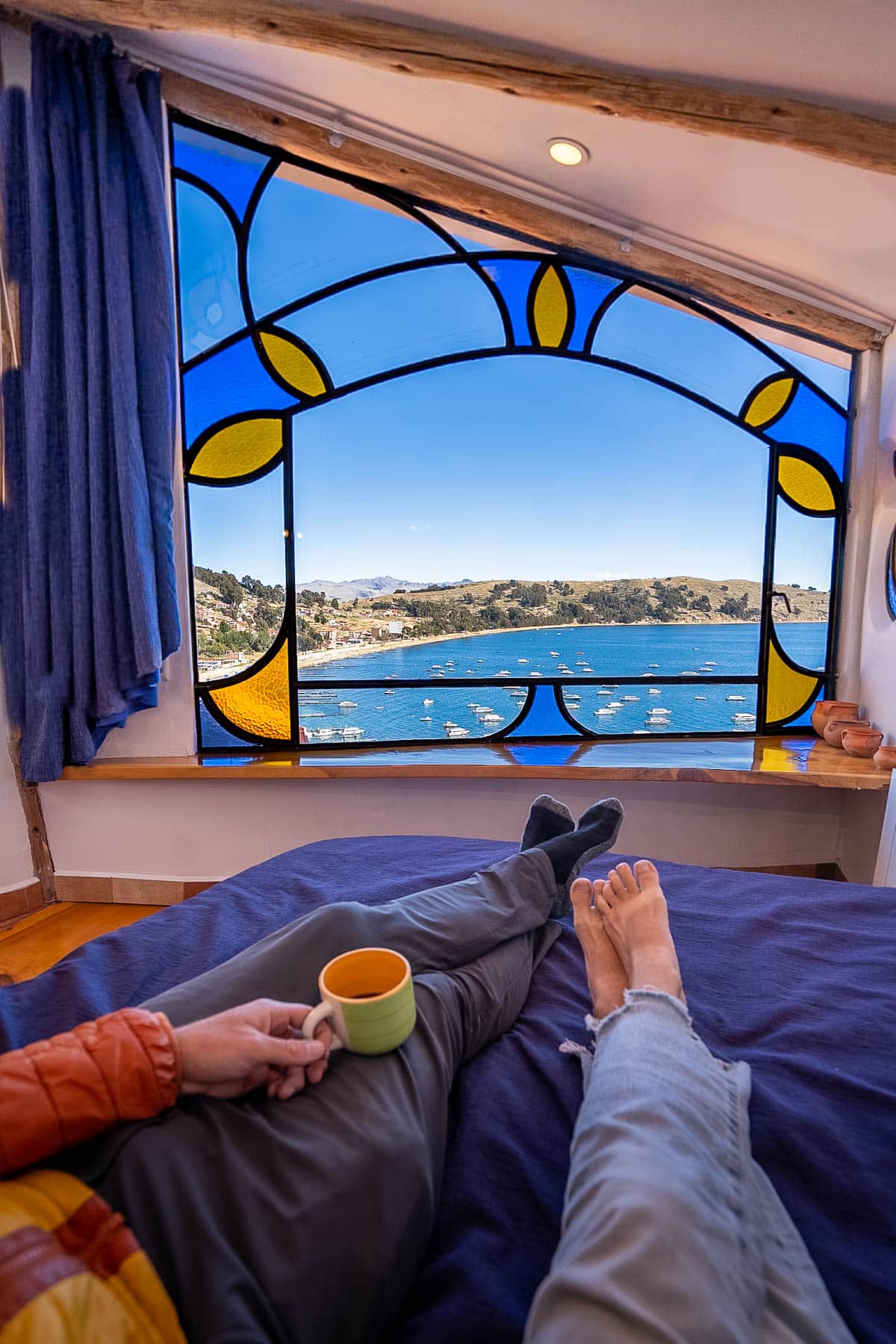 Couple's feet sitting on a bed with a window that overlooks Lake Titicaca at Hostal las Olas in Copacabana, Bolivia
