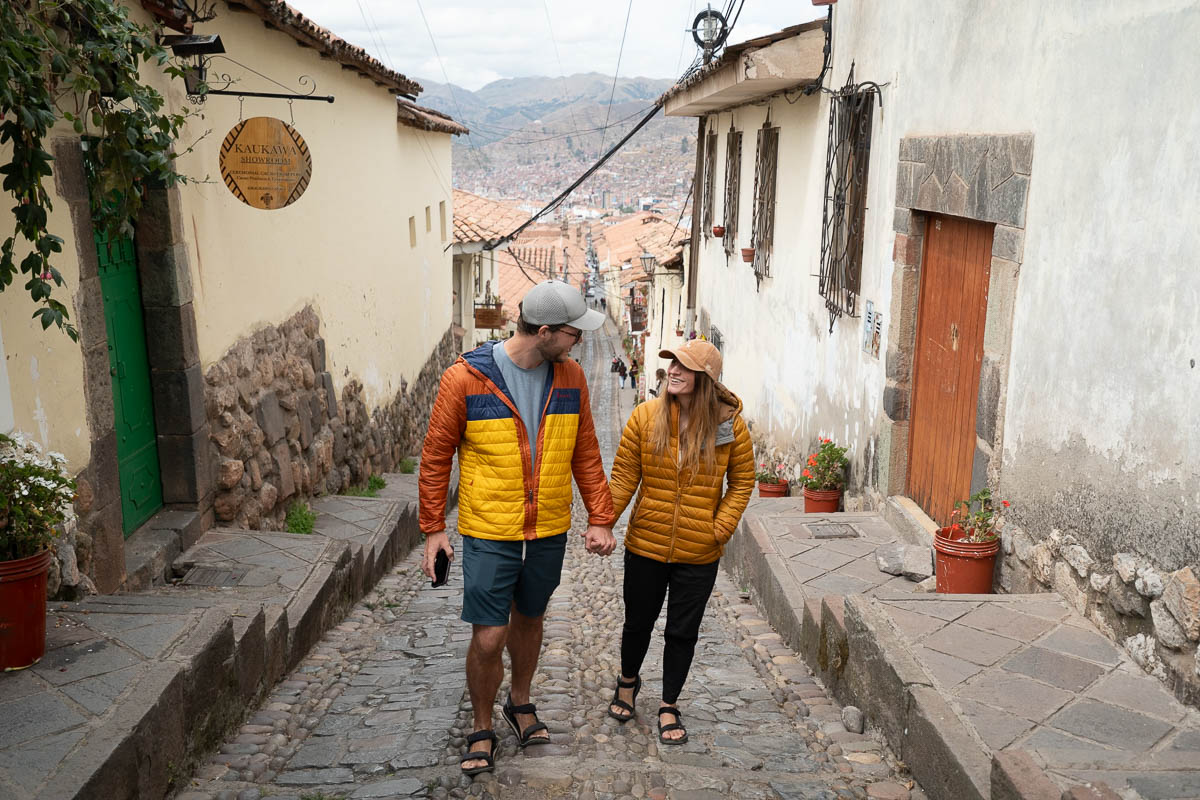 Couple holding hands walking down a cobblestone street in the San Blas neighborhood with mountains in the background in Cusco, Peru
