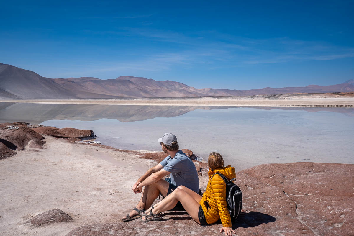 Couple sitting on red rocks at Piedras Rojas with a lagoon and mountains in the background in the Atacama Desert, Chile