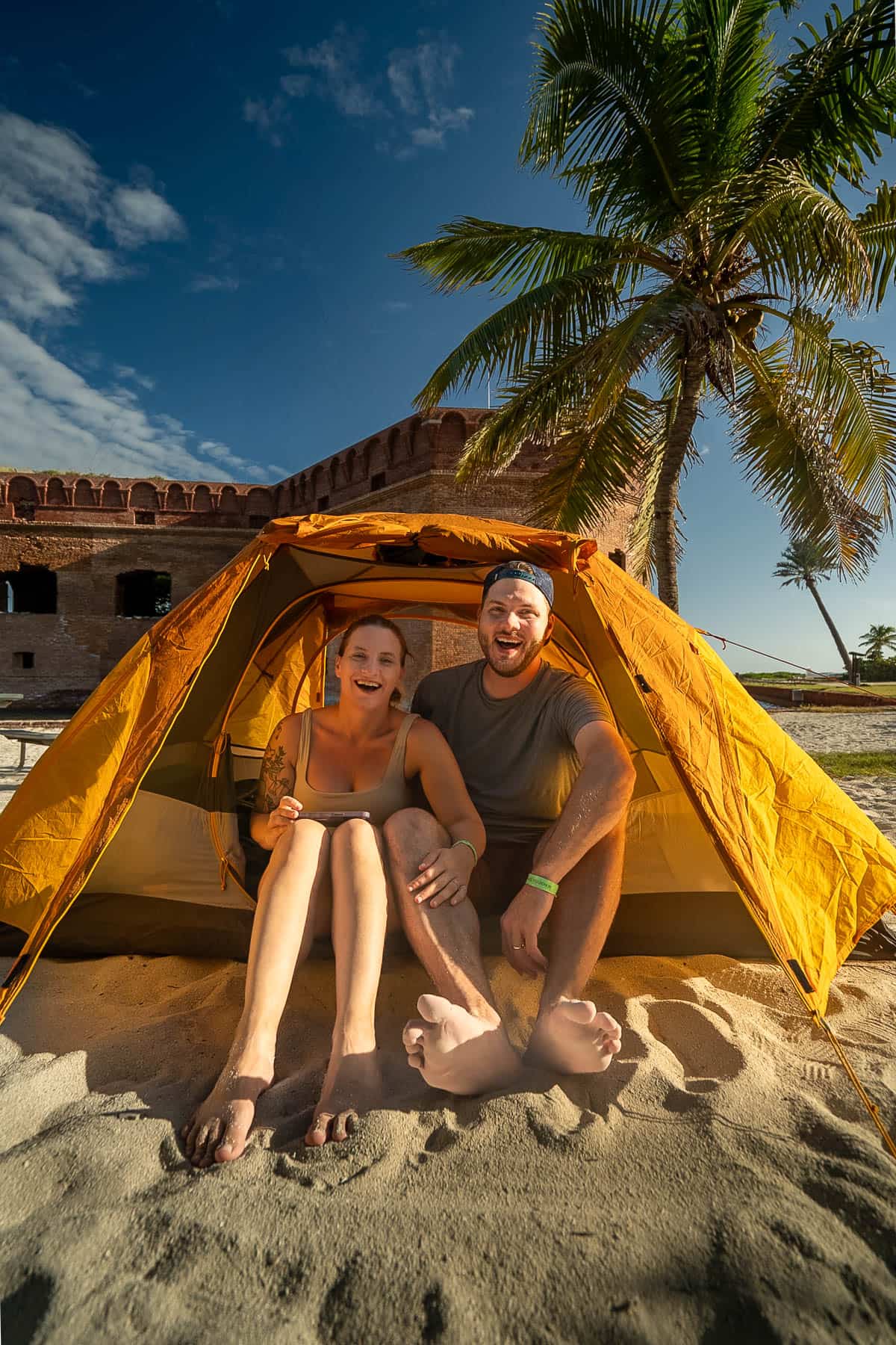 Smiling couple sitting in a tent with a coconut tree and Fort Jefferson in the background in Dry Tortugas National Park in Florida