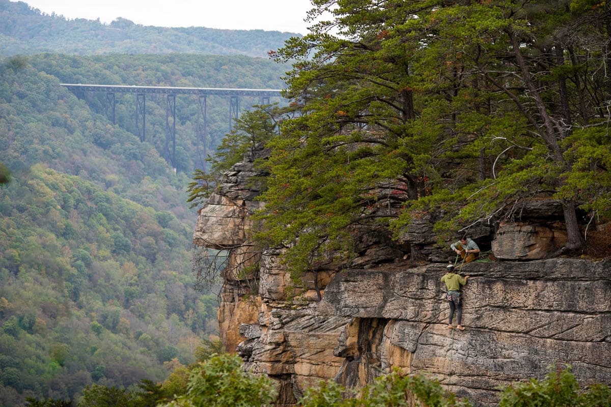 Two rock climbers climbing down sandstone cliff with the New River Gorge National Park with the New River Gorge bridge in the background along the Endless Wall Trail in New River Gorge National Park in West Virginia