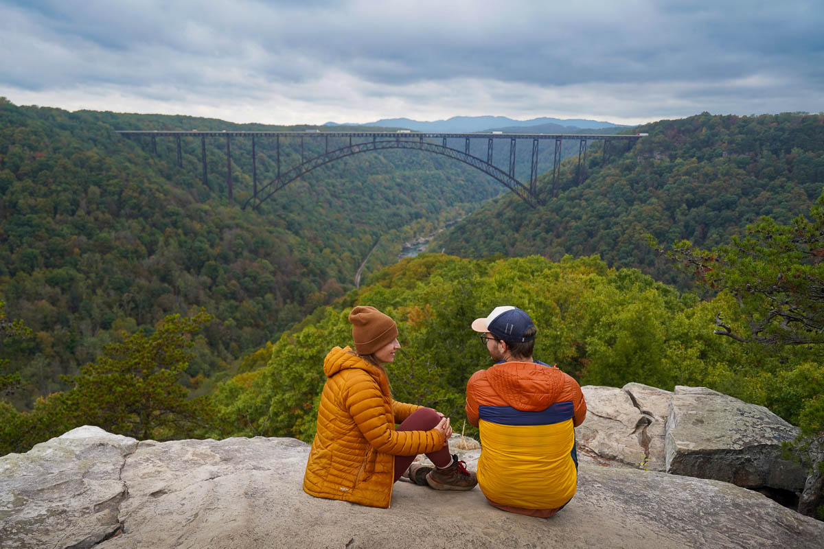 Couple sitting on rocky outcropping with the New River Gorge Bridge in the background along the Long View Trail in New River Gorge National Park in West Virginia