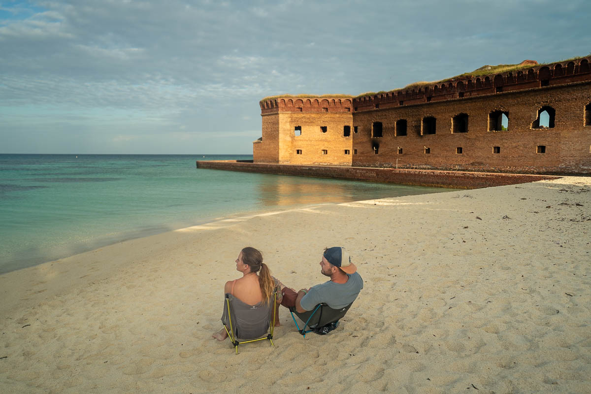 Couple sitting in camping chairs on a white sandy beach with Fort Jefferson in the background at Dry Tortugas National Park near Key West, Florida