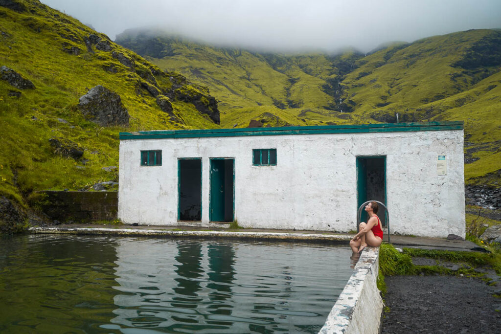 Woman sitting on the edge of a concrete lip of the Seljavallalaug Swimming Pool with a green cliffside in the background in Iceland