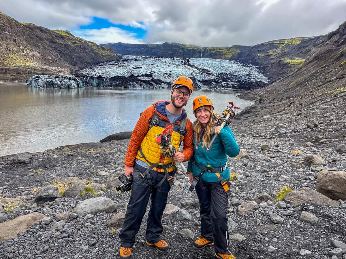 Couple wearing a helmet, harness, and ice axe with the Sólheimajökull Glacier and mossy mountains in the background in Iceland