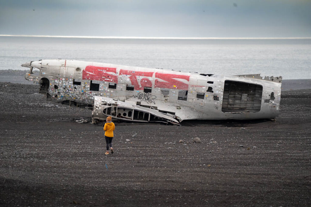 Woman walking towards the Solheimasandur Plane Wreck on a black sand beach with Atlantic Ocean in the background in Iceland