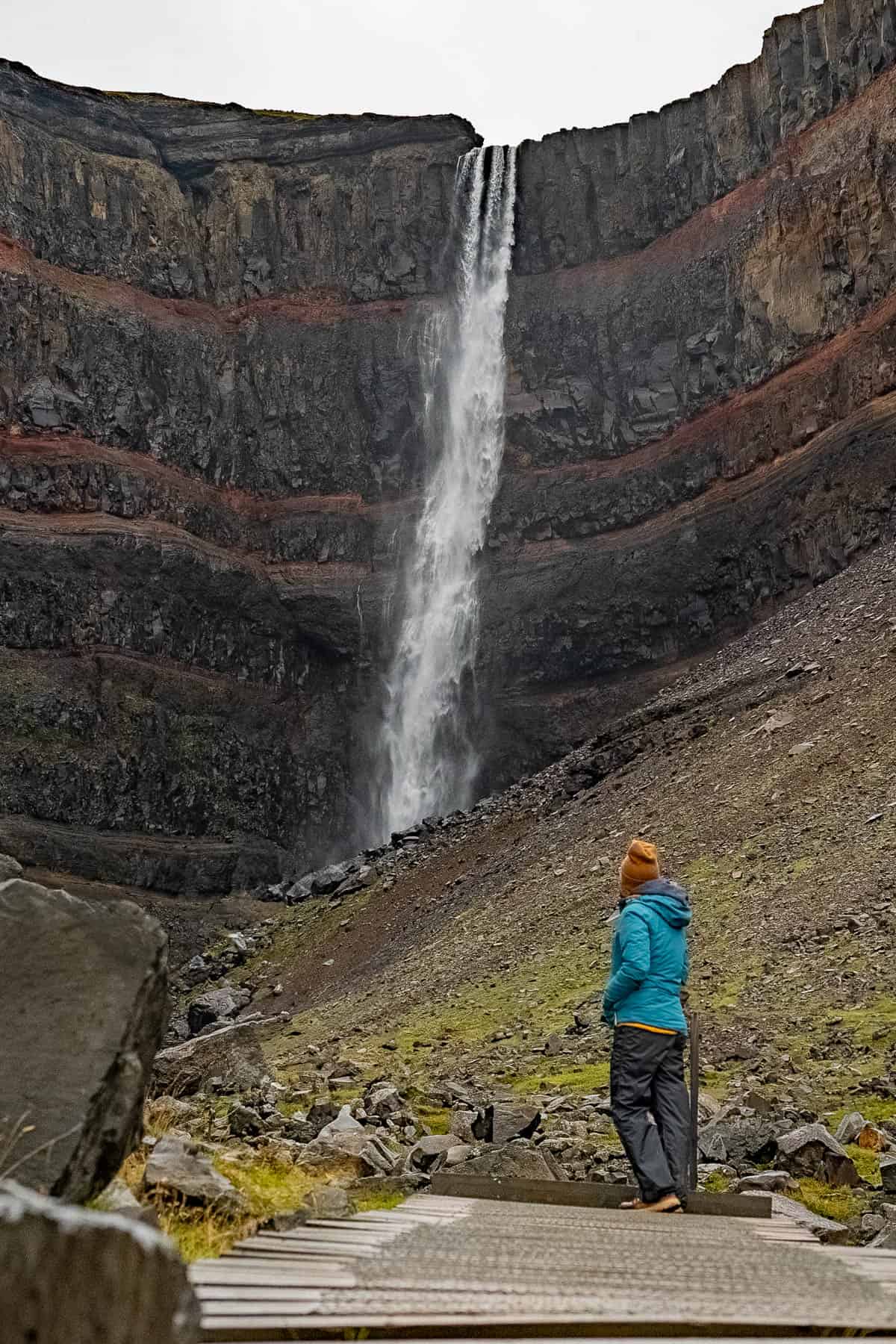 Woman standing at the end of a wooden boardwalk with a basalt cliffside with red volcanic rocks with Hengifoss Waterfall in Iceland