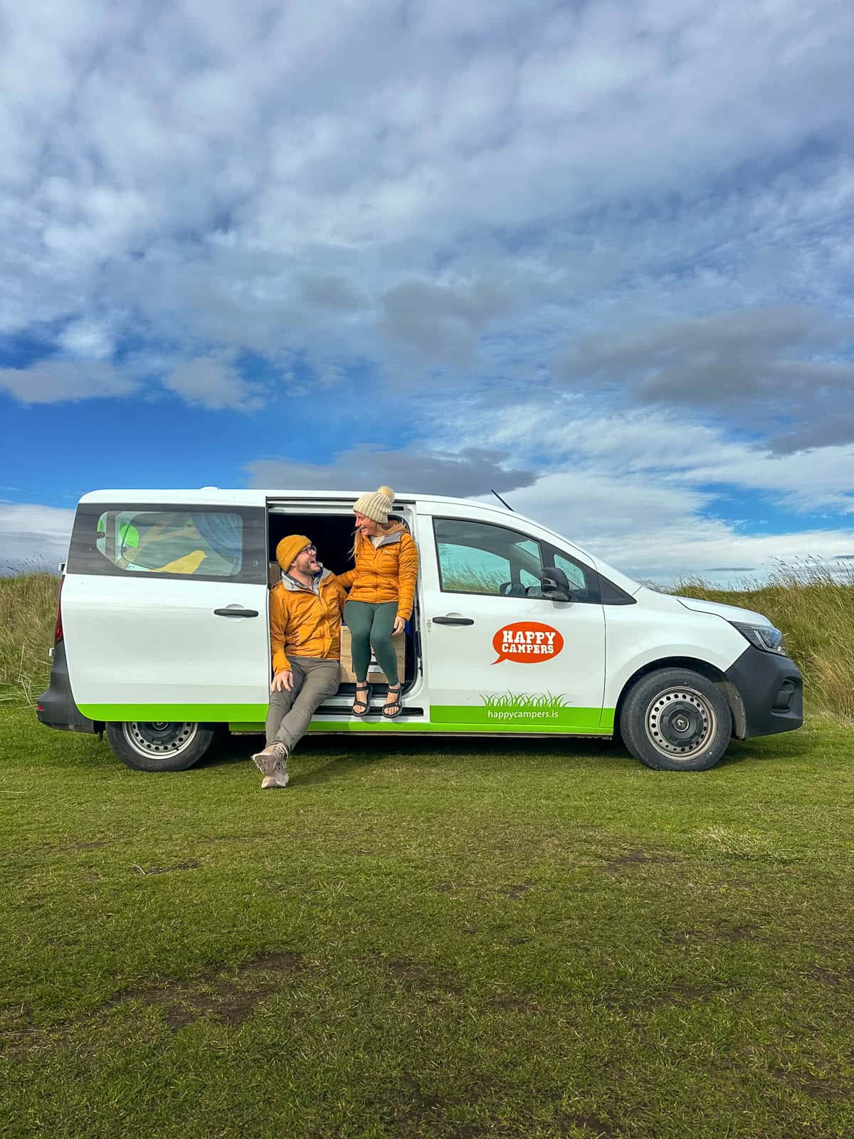 Couple smiling in the open door in a Happy Campers campervan, parked in a grassy field in Iceland