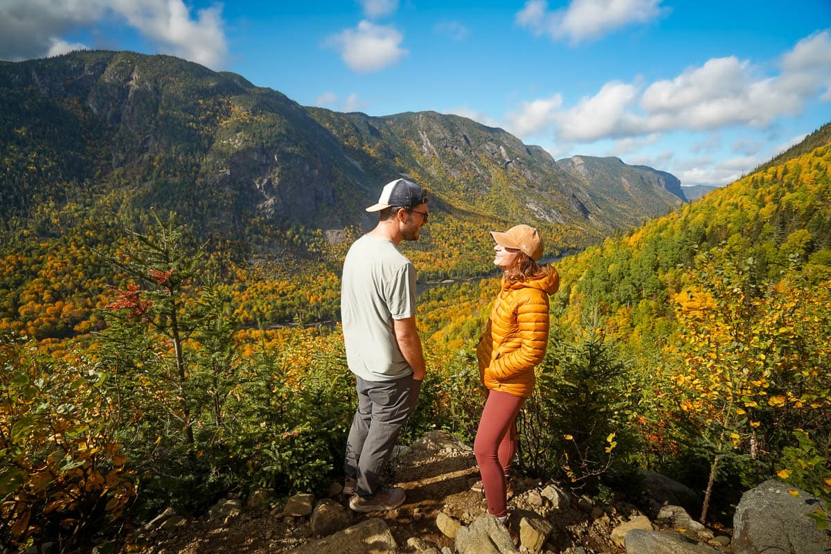 Couple standing at a viewpoint with a valley in the background with fall foliage on the hillsides along the Acropole des Draveurs trail in Hautes Gorges de la Riviere Malbaie in Quebec, Canada