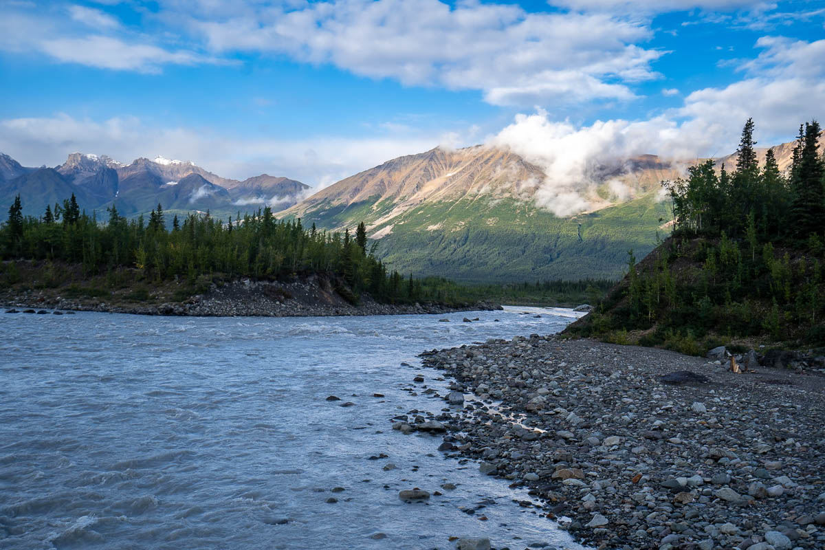 Kennicott River with rocky shoreline surrounded by pine trees and mountains in the background outside of McCarthy, Alaska