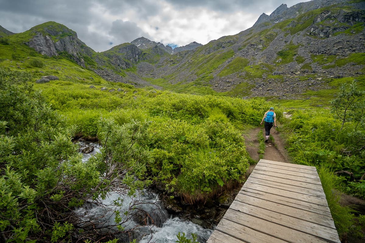 Woman crossing a wooden bridge with rugged mountains in the background along the Reed Lakes Trail in Hatcher Pass, Alaska