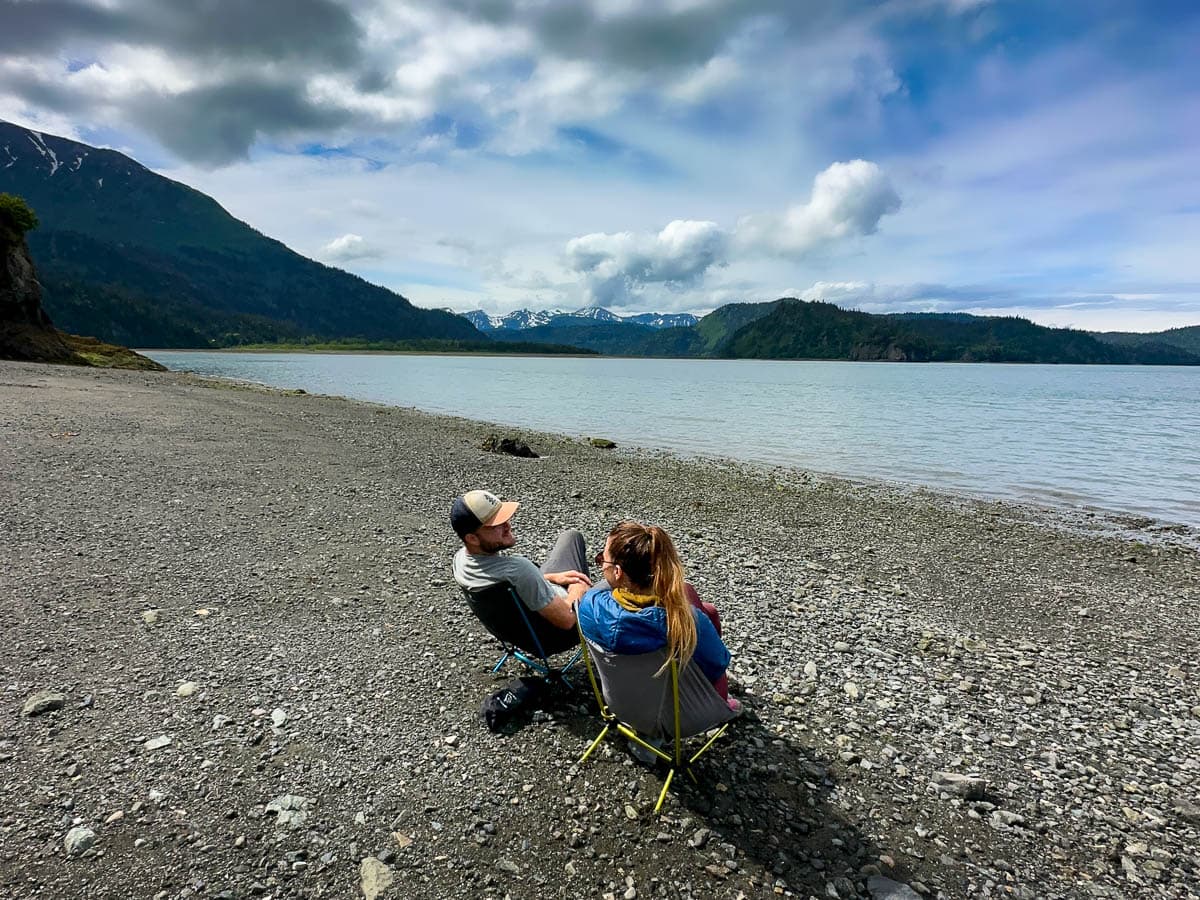 Couple sitting in camping chairs along a rocky beach near the Saddle trailhead with mountains in the background in Kachemak Bay State Park, Alaska