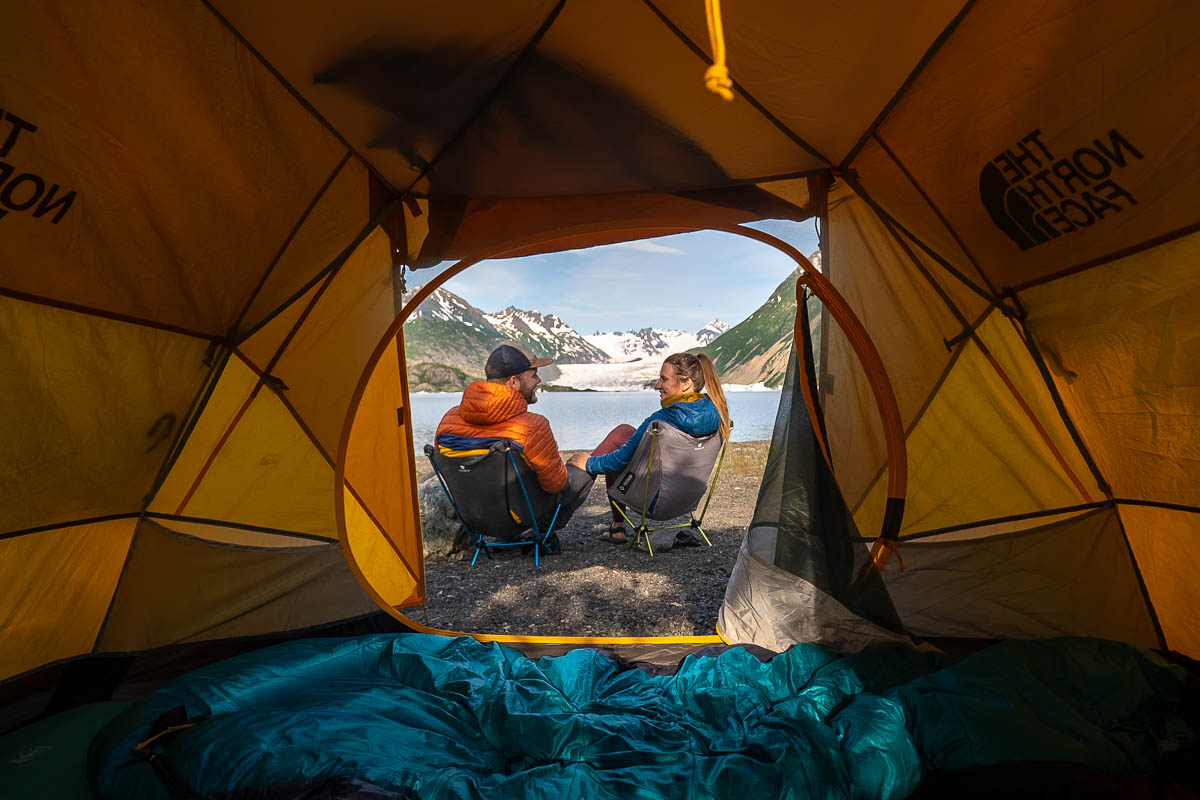 Couple sitting camping chairs out of a tent opening with the Grewingk Glacier Lake in the background in Kachemak Bay State Park, Alaska