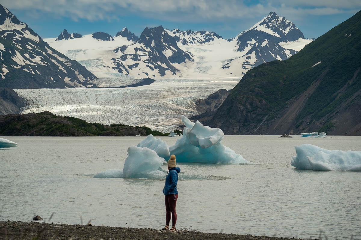 Woman standing along a shoreline with icebergs in the Grewingk Glacier Lake in the background in Kachemak Bay State Park, Alaska