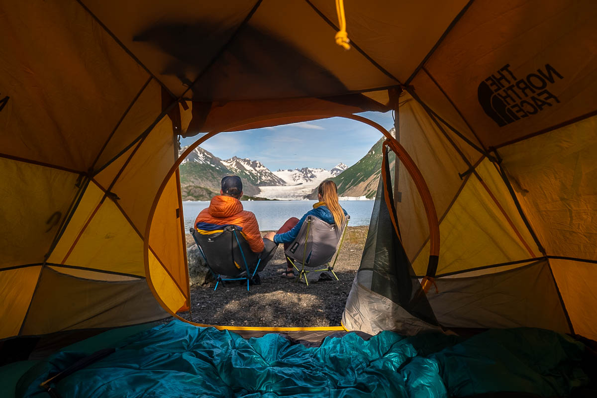 Couple sitting in a camping chair outside of their tent with Grewingk Glacier Lake in Kachemak Bay State Park, Alaska