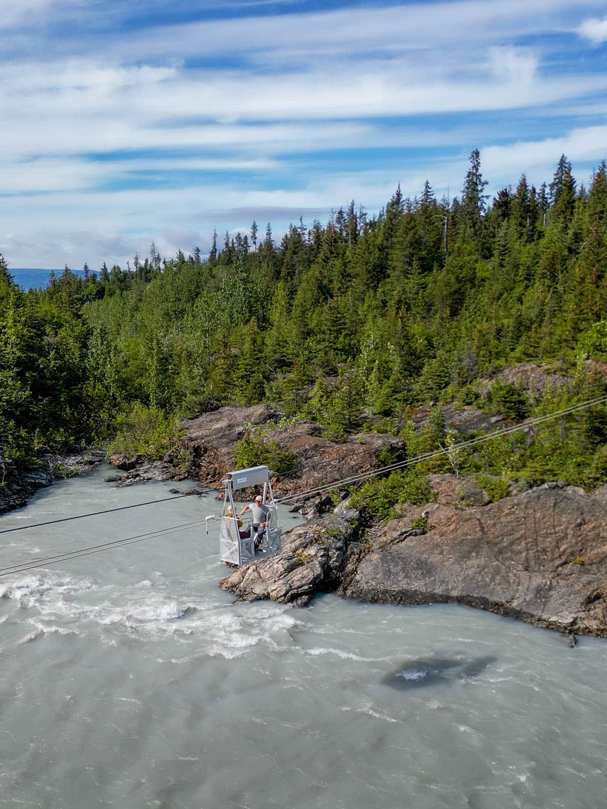 Couple sitting in a tram over the Grewingk Creek with pine trees in the background along the Emerald Lake Trail in Kachemak Bay State Park, Alaska