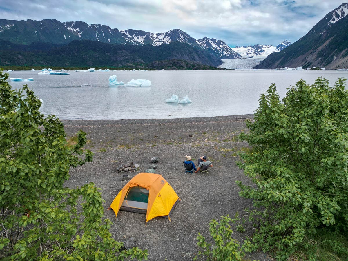 Couple holding hands while sitting in camping chairs next to their tent with the Grewingk Glacier Lake in the background in Kachemak Bay State Park, Alaska