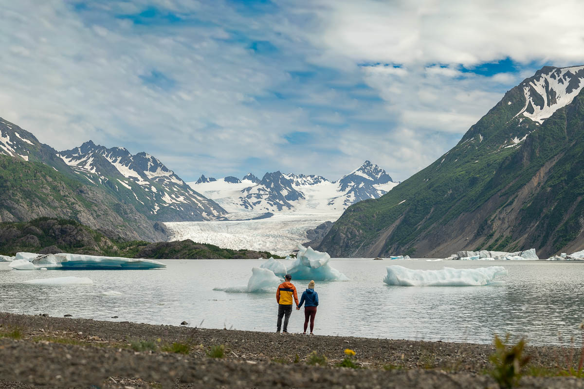 Couple holding hands along a rocky beach in front of Grewingk Glacier Lake with mountains in the background in Kachemak Bay State Park, Alaska