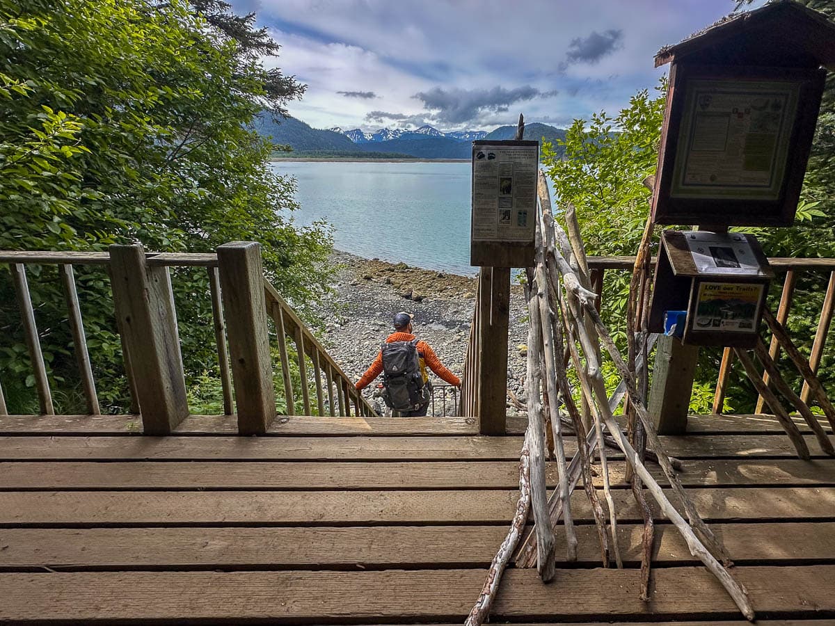 Man walking down wooden stairs to the Saddle trailhead with snowcapped mountains in the background in Kachemak Bay State Park, Alaska