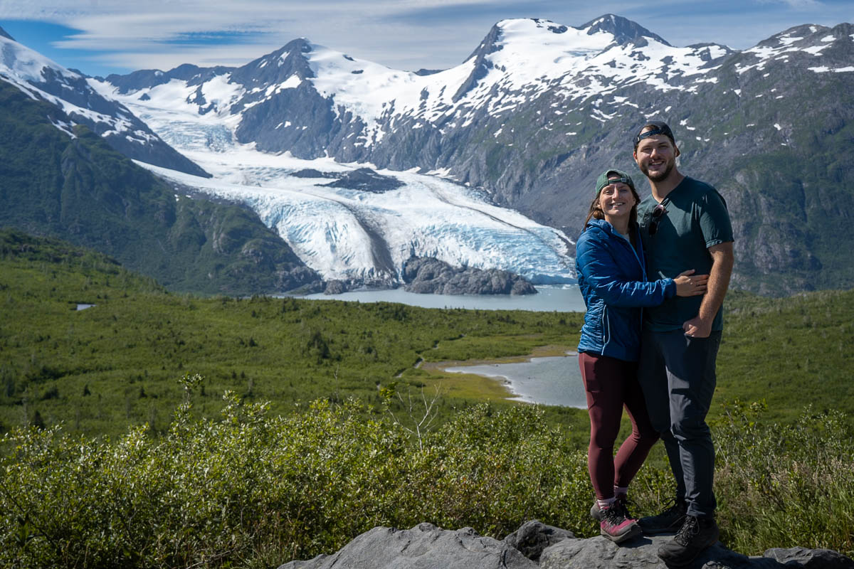 Couple smiling on top of a rocky outcropping with Portage Glacier and mountains in the background along the Portage Pass Trail in Whittier, Alaska