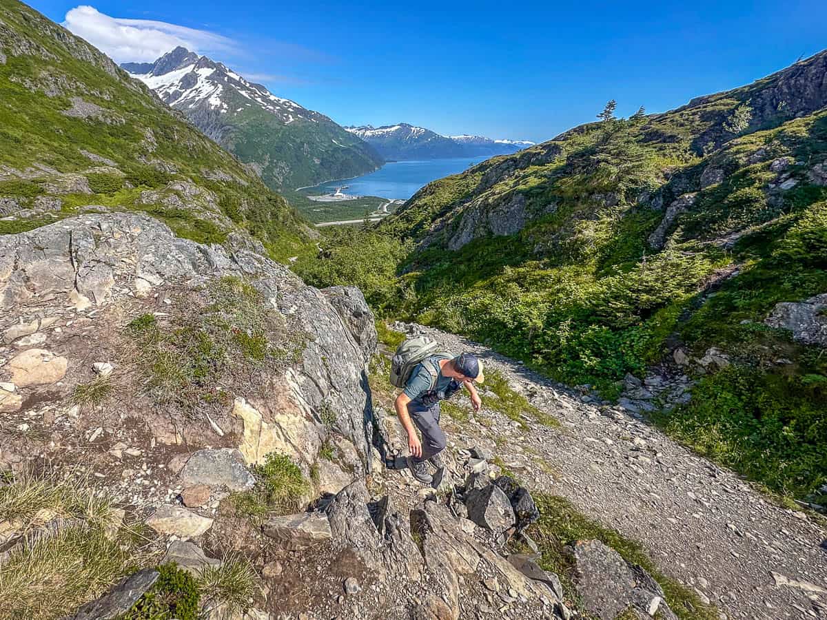 Man climbing down a rocky outcropping along the Portage Pass Trail with mountains and the Prince William Sound in the background in Whittier, Alaska