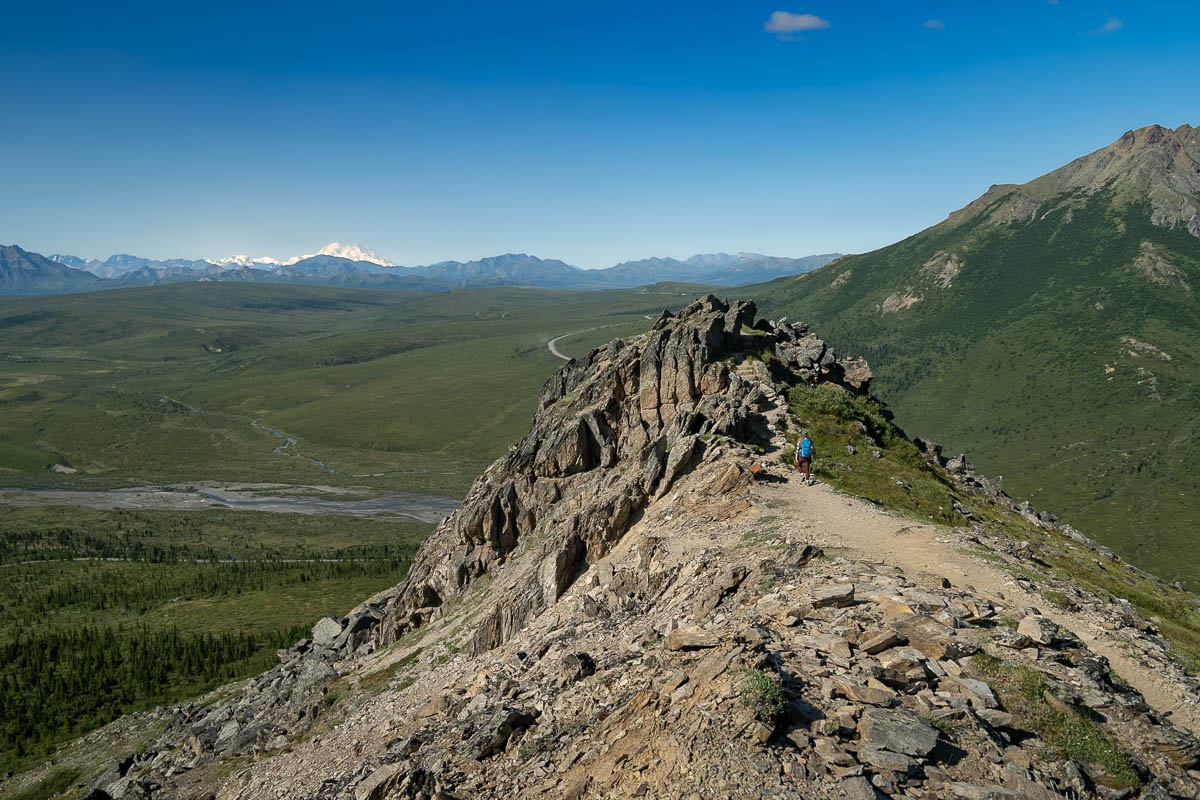 Woman hiking towards rocky formation with Mount Denali in the background along the Savage Alpine Trail in Denali National Park, Alaska