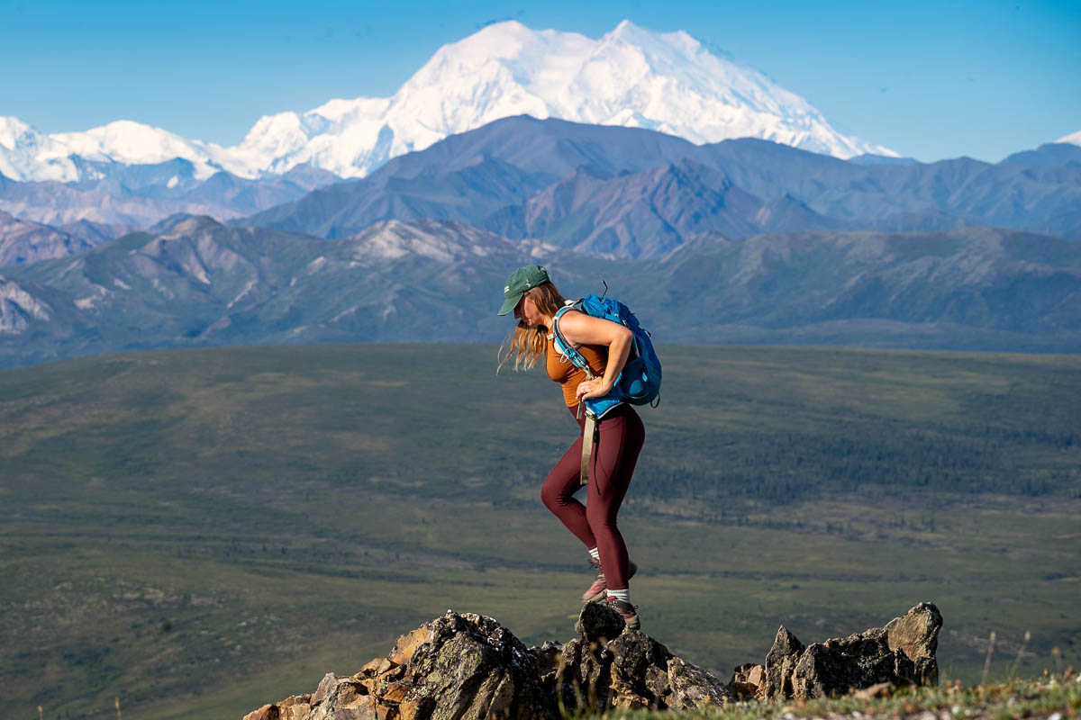 Woman hiking up a rocky outcropping with Mount Denali and other mountains in the background along the Savage Alpine Trail in Denali National Park, Alaska